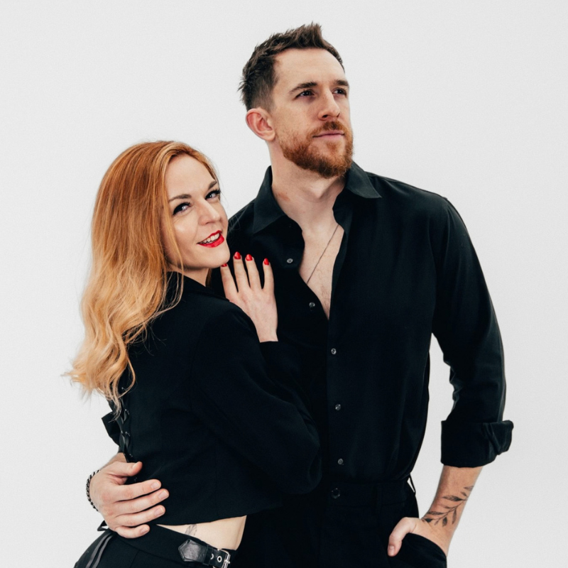 A man and a woman posing together against a plain light gray studio background. The man wears a black button-up shirt, and the woman wears a black cropped top and black bottoms.