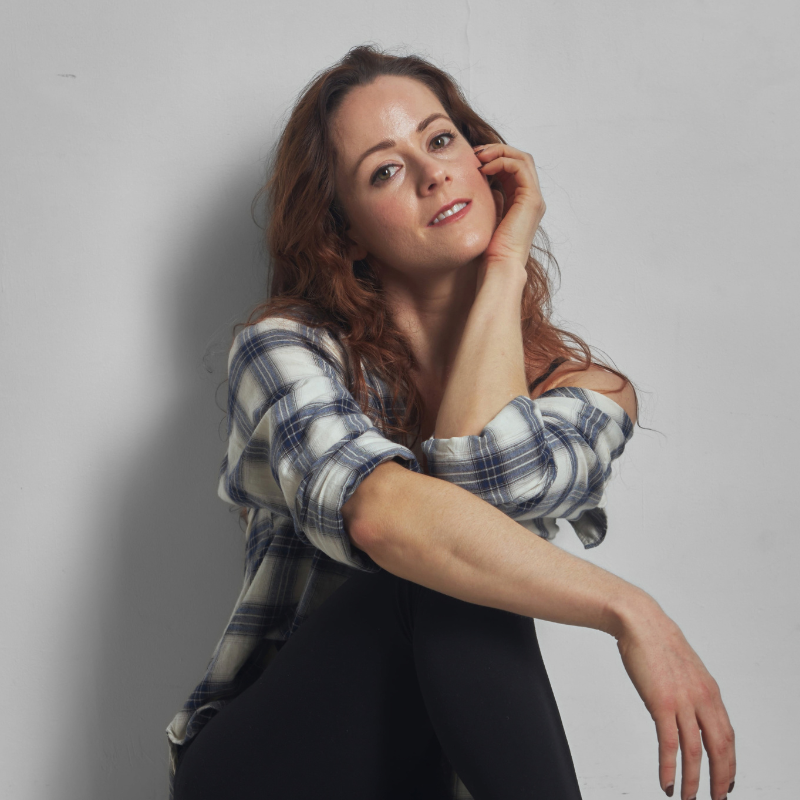 A woman with long wavy auburn hair leans against a light gray wall, wearing a blue, white, and gray plaid flannel shirt over black pants.