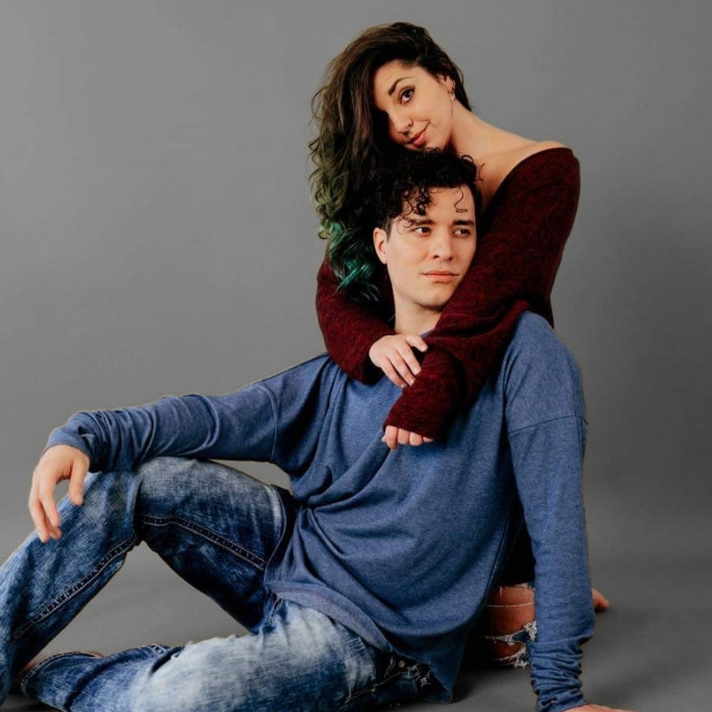 A man and a woman posing together against a solid gray studio background. The man is sitting and wearing a blue long-sleeve shirt and jeans, while the woman sits behind him wearing a dark red off-the-shoulder knit sweater.