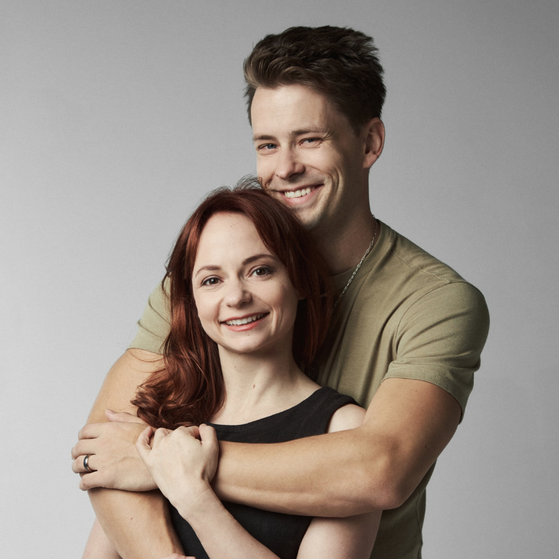 A smiling man in an olive green t-shirt and a woman with brown hair in a black top, hugging against a solid gray studio background.