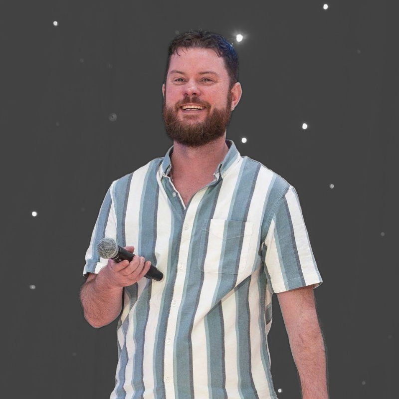 A smiling man with a beard holding a microphone, wearing a short-sleeve button-up shirt with vertical light blue and white stripes, set against a dark gray background with small white star-like specks.