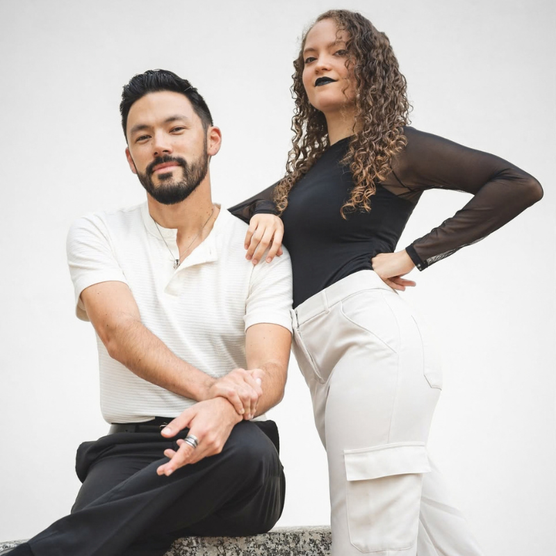 A man and a woman posing together against a plain light background. The man is seated wearing a white short-sleeve polo shirt and black pants, while the woman stands wearing a black sheer-sleeve long-sleeve top and white pants.