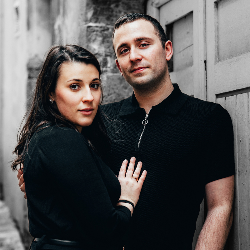 A man and a woman posing together in a narrow alleyway. The man is wearing a black short-sleeve polo shirt with a zipper collar, and the woman is wearing a black long-sleeve top.