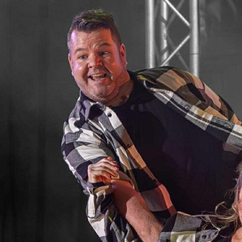 A man wearing a black t-shirt and a brown, black, and white plaid flannel shirt, captured mid-gesture on a stage with lighting and truss rods visible in the background.