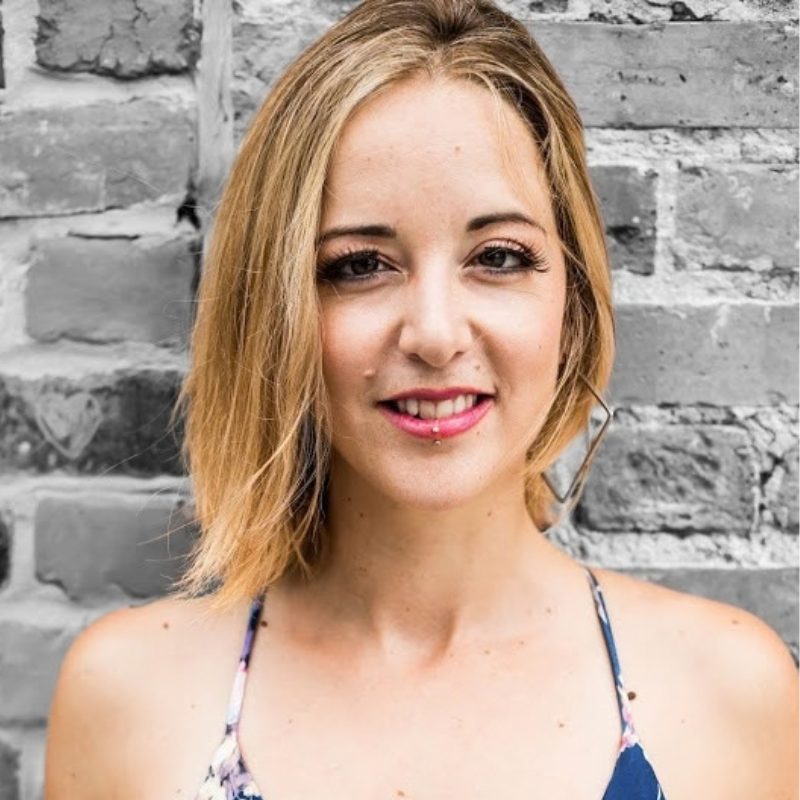 A smiling blonde woman with a small lip piercing standing in front of a gray brick wall.